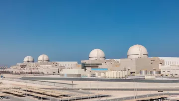 L'image montre une centrale nucléaire moderne avec plusieurs grands dômes blancs sur le toit. La structure est entourée d'un paysage clair, avec un ciel bleu sans nuages. À l'avant-plan, on peut voir des bâtiments de différentes tailles, ainsi qu'une zone de stationnement. L'ensemble de la scène dégage une impression de technologie avancée et de robustesse.