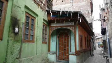 L'image montre une rue étroite et pittoresque dans une vieille ville, avec des maisons en bois et en pierre. On peut voir des façades colorées, notamment des murs verts et oranges, ainsi que des fenêtres à volets en bois. Les bâtiments ont un style architectural traditionnel, avec des détails ornementaux visibles. Des fils sont tendus entre les maisons, probablement pour des décorations ou des lumières. L'atmosphère évoque un sentiment de nostalgie et de charme ancien.