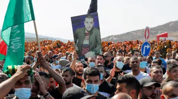الصورة تُظهر مجموعة من المتظاهرين في تجمع حاشد، يحمل بعضهم لافتات وأعلام. وسط الحشد، يوجد شخص يحمل صورة لرجلٍ يبدو أنه شخصية مهمة أو رمزية بالنسبة لهم. يرتدي الكثير من المتظاهرين الكمامات، ويبدو أن المظاهرة تتم في منطقة مفتوحة، مع خلفية تضم تلالًا وأشجار. تُعبر الأجواء عن الحماس والتجمع الجماهيري.