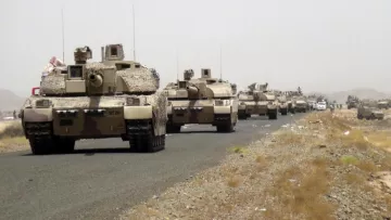 L'image montre une colonne de véhicules militaires, probablement des chars, se déplaçant sur une route désertique. Les véhicules sont camouflés dans des tons de beige et de marron, adaptés à l'environnement. On peut voir un paysage aride en arrière-plan, avec de la terre et quelques collines. L'atmosphère générale semble être celle d'un déploiement militaire.