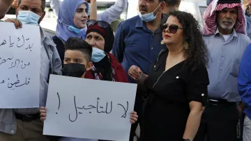En la imagen se puede ver a un grupo de personas reunidas, algunas de las cuales sostienen carteles. La mayoría llevan mascarillas. Entre las personas, un niño sostiene un cartel que tiene un mensaje. Se observa una variedad de expresiones en sus rostros, reflejando una atmósfera de protesta o manifestación. La vestimenta de los participantes es variada, con algunas personas usando prendas tradicionales. El ambiente sugiere una situación de movilización social.