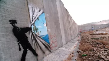 L'image montre un mur de béton, probablement un mur de séparation ou de sécurité, sur lequel est peint un graffiti représentant un policier. Ce dernier semble tirer un rideau pour révéler un paysage idyllique, avec des palmiers et un ciel bleu, symbolisant un contraste entre la réalité de l'environnement urbain et un espace rêvé ou libre. Sur le mur, on peut également voir des inscriptions en arabe. L'ensemble de l'image peut évoquer des thèmes de restriction, d'espoir et de liberté.