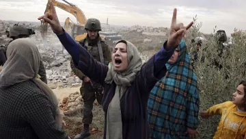 L'immagine mostra un gruppo di persone in una situazione di grande tensione emotiva. Una donna al centro, con un velo, alza le braccia in segno di protesta o di disperazione. Il suo volto esprime forte emozione. Intorno a lei, ci sono altre donne e un bambino, tutte visibilmente colpite dalla situazione. Sullo sfondo, si intravede un escavatore e una presenza militare, suggerendo che si sta svolgendo un'operazione di demolizione o sgombero. Il contesto sembra essere caratterizzato da conflitto e tensioni sociali.