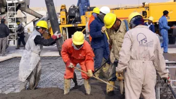 الصورة تظهر مجموعة من العمال في موقع بناء وهم يعملون على صب الخرسانة. يرتدي العمال خوذات لحماية الرأس وأزياؤهم متنوعة الألوان، حيث يركز البعض على تجريف الخرسانة باستخدام معدات يدوية. تظهر في الخلفية شاحنة وأدوات أخرى تتعلق بأعمال البناء، مما يدل على نشاط مكثف في الموقع.