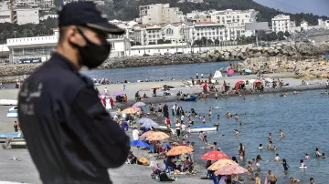 تظهر الصورة شاطئاً مزدحماً حيث يتواجد العديد من الأشخاص الذين يستمتعون بالسباحة والاسترخاء تحت الشمس. يوجد في الصورة مجموعة من المظلات الملونة التي توفر الظل للزوار. في المقدمة، هناك رجل أمن يرتدي كمامة وبدلة رسمية، يراقب الأوضاع. يظهر في الخلفية مجموعة من المباني والجبال، مما يضفي طابعاً جميلاً على المشهد.