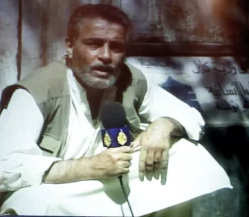 L'image montre un homme assis, tenant un micro et portant une tenue traditionnelle. Il a une barbe grise et semble parler avec intensité, probablement en train de faire un reportage. En arrière-plan, il y a un panneau ou une affiche qui n'est pas clairement visible. L'éclairage de l'image semble un peu flou, ajoutant une ambiance particulière à la scène.