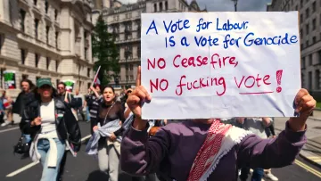 L'image montre une manifestation où des personnes défilent dans la rue, tenant des pancartes. Au premier plan, une personne tient une grande affiche avec un message fort. La pancarte indique que voter pour un certain parti est associé à des accusations graves, et elle appelle à ne pas voter sans un cessez-le-feu. Les manifestants semblent passionnés et engagés, exprimant leur désaccord par des slogans. L'ambiance est empreinte de tension et d'émotion.
