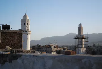 L'image montre une vue panoramique d'une ville avec des minarets distinctifs. On peut voir deux tours blanches caractéristiques, l'une étant en cours de restauration, entourées d'un paysage montagneux en arrière-plan. Le ciel est clair, indiquant une ambiance paisible au crépuscule. Au loin, plusieurs autres structures architecturales, probablement des mosquées, se dessinent à l'horizon, contribuant à l'atmosphère culturelle et historique de la scène.