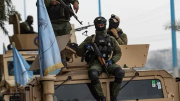 L'image montre des soldats en uniforme, armés et équipés de casques, se trouvant sur un véhicule militaire. L'un des soldats est assis sur le toit du véhicule, tenant une arme, tandis que d'autres soldats sont debout à proximité. On peut également voir un drapeau, probablement lié à une mission de maintien de la paix. L'environnement semble urbain, avec des éléments de la ville en arrière-plan.