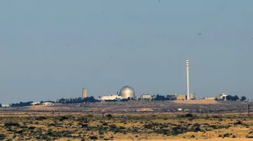 الصورة تظهر مشهداً لبيئة طبيعية صحراوية مع مبانٍ تظهر في الخلفية. يبدو أن هناك مرفقاً يحتوي على قبة كبيرة، ومن المحتمل أن يكون هذا المرفق مركزاً علمياً أو مفاعلاً نووياً. كما يُلاحظ وجود أعمدة مدخنة تشير إلى أنشطة صناعية. السماء صافية واللون العام للصورة يميل إلى الألوان الترابية.