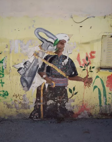 L'image montre une fresque sur un mur. On y voit un personnage portant un costume traditionnel, avec un air déterminé. Ce personnage tient une plante dans une main, symbolisant la vie et la nature. Sur son dos, on peut apercevoir une sorte d'outil ou d'équipement qui pourrait symboliser le travail ou un engagement envers la terre. Le fond est de couleur jaune, ce qui attire l'œil et met en avant le contraste avec les couleurs du personnage. Des inscriptions et des graffitis en arrière-plan ajoutent du caractère à l'œuvre.
