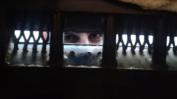 L'image montre un regard émergeant à travers une grille ou une ouverture sombre. Les yeux de la personne sont bien visibles, tandis que le reste de son visage est caché par l'ombre ou la structure. Les bords de l'ouverture semblent usés, ajoutant une atmosphère de confinement ou de mystère à la scène.