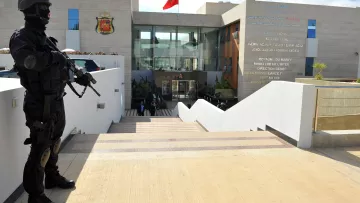 The image depicts a heavily armed police officer or security personnel standing at attention outside a building. The officer is dressed in black tactical gear and is holding a firearm. In the background, there is a modern structure with a large entrance, and the Moroccan flag is prominently displayed on top. The building appears to be a government facility, likely related to security or law enforcement, as indicated by the presence of the armed guard. The setting features stairs leading up to the entrance, with a clean, contemporary design.