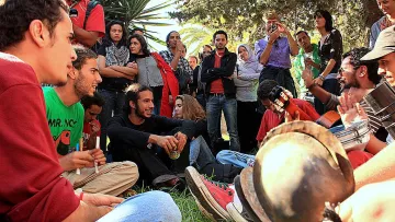 L'image montre un groupe de personnes rassemblées dans un environnement extérieur, probablement dans un parc ou un jardin. Des jeunes, assis sur l'herbe, semblent participer à une activité musicale, avec des instruments comme une guitare et des percussions. Certains d'entre eux sont engagés dans une discussion animée. En arrière-plan, d'autres personnes regardent ou écoutent, créant une atmosphère conviviale et collective. Les expressions des participants varient, allant de la concentration à l'amusement.