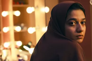 L'image montre une jeune femme avec un regard pensif, portant un hijab. Elle est positionnée de manière à ce que son visage soit tourné légèrement vers la caméra, tandis qu'en arrière-plan, on aperçoit un miroir entouré de lumières douces, créant une ambiance chaleureuse et intime. Son expression reflète une certaine introspection, ajoutant une profondeur émotionnelle à la scène.