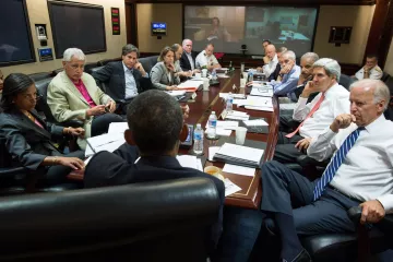 L'image montre une réunion importante autour d'une grande table. Plusieurs personnes, des hommes et des femmes, sont assis, engagés dans une discussion sérieuse. À l'arrière-plan, on peut voir un écran où se tient une vidéoconférence. Les participants semblent être des responsables et des conseillers, probablement dans un contexte gouvernemental ou militaire. L'ambiance générale est concentrée, avec des documents étalés sur la table et des bouteilles d'eau à disposition.