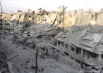 L'image montre un paysage urbain fortement endommagé, avec des bâtiments effondrés et en ruines. Les façades des immeubles sont détruites, laissant entrevoir des débris et des structures instables. Le sol est recouvert de débris et de poussière, témoignant de la violence des destructions. La lumière du jour éclaire la scène, accentuant le contraste entre les ruines et le ciel. Cette image évoque un environnement de guerre et de crise humanitaire.