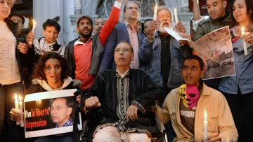 L'immagine mostra un gruppo di persone radunate in un ambiente urbano, apparentemente in un momento di protesta o di vigilanza. Alcuni partecipanti tengono in mano candele accese, mentre altri mostrano cartelli e manifesti con slogan che richiamano il tema della libertà di espressione. Tra le persone c'è un uomo in sedia a rotelle, che sembra essere al centro dell'attenzione. L'atmosfera è di solidarietà e impegno per i diritti civili.