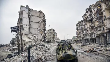 تصویر نشان‌دهندهٔ یک منطقهٔ ویران شده است که در آن چند ساختمان تخریب شده و در حال فرو ریختن به نظر می‌رسند. در این صحنه، یک وسیله نقلیه نظامی در حال حرکت است و تعدادی سرباز در آن نشسته‌اند. بقیهٔ محیط شامل زباله‌ها و نشانه‌های ویرانی است که به وضعیت بحرانی منطقه اشاره دارد. هوا ابری و تیره به نظر می‌رسد و فضایی خالی و ساکت را ایجاد کرده است.