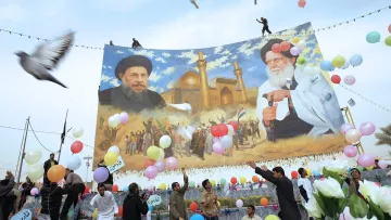 L'image montre une scène festive en extérieur avec un grand panneau représentant deux figures, probablement des leaders ou des personnalités importantes. Au-dessous de ce panneau, des gens célèbrent, tenant des ballons colorés et des fleurs. Certaines personnes semblent danser ou exprimer leur joie. En arrière-plan, il y a un décor architectural qui semble être un bâtiment religieux, avec des minarets visibles. Des colombes volent également dans le ciel, ajoutant une dimension symbolique à l'événement.