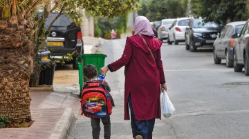 تظهر في الصورة امرأة ترتدي حجابًا وتلبس معطفًا طويلًا، تمشي مع طفل صغير يحمل حقيبة مدرسية على ظهره. يبدو الطفل سعيدًا وهو يمسك بيد والدته. الشارع محاط بالأشجار والمنازل، وهناك سيارات متوقفة على الجانبين. الأجواء تبدو هادئة وتعكس لحظة يومية من حياة الأسرة.