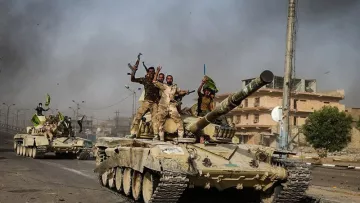 الصورة تظهر مجموعة من الجنود على دبابات أثناء التحرك في منطقة حضرية، حيث يرفع بعضهم الأعلام ويعبرون عن الفرح والنصر. الخلفية تحتوي على مباني مدمرة وأجواء مليئة بالدخان، مما يشير إلى وجود نشاط عسكري. الدبابات تبدو قديمة الطراز، ويعكس المشهد التوترات والصراعات في المنطقة.