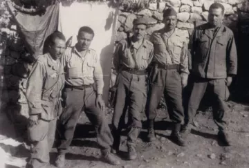 L'image présente un groupe de cinq hommes en uniforme militaire, debout ensemble devant un mur en pierres. Les hommes semblent être d'origine variée, portant des vêtements similaires, avec des pantalons et des chemises. Deux d'entre eux tiennent des armes à feu, et leur expression dégage un sentiment de camaraderie et de détermination. En arrière-plan, on aperçoit un drap suspendu, ce qui suggère qu'ils se trouvent dans un camp ou un endroit de rassemblement. L'atmosphère est sérieuse, typique des contextes militaires.