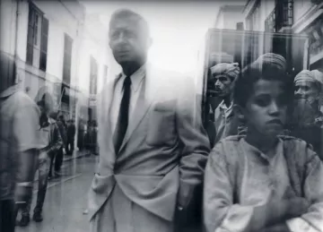 L'image semble capturer une scène de rue en noir et blanc, où se mêlent des figures humaines. Au premier plan, un homme en costume clair se tient debout, semblant légèrement détaché du reste de la foule. En arrière-plan, on aperçoit plusieurs personnes, probablement des habitants, qui regardent vers la caméra. L'ambiance est floue, ce qui donne une impression de mouvement et de vie dynamique dans la rue. Les vêtements traditionnels des personnes en arrière-plan contrastent avec le style moderne de l'homme au premier plan, créant une image intéressante et expressive.