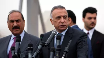 تظهر الصورة مجموعة من الرجال أثناء مؤتمر صحفي. في المقدمة يوجد رجل يرتدي بدلة رسمية، ويبدو جادًا وهو يتحدث أو يستعد للإدلاء ببيان. خلفه، يوجد رجل آخر يرتدي بدلة أيضًا، يبدو أنه يستمع أو يشارك في الحوار. هناك عدد من الميكروفونات أمامهم، مما يوحي بأنهم في موقف يتطلب إلقاء الخطابات أو التصريحات الإعلامية. الجو العام يبدو رسميًا، وقد تكون هناك بعض الضبابية في الخلفية.