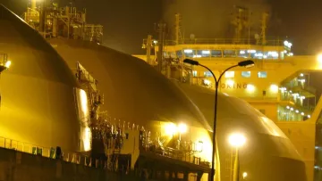 L'image montre une scène nocturne avec de grands réservoirs de stockage, probablement liés à l'industrie du gaz naturel. Les réservoirs ont des formes arrondies et sont éclairés par des lumières jaunes, créant une ambiance industrielle. En arrière-plan, on peut voir un navire, équipé de dispositifs de chargement ou de déchargement, suggérant une activité portuaire. La combinaison de l'illuminations et des structures métalliques donne une impression de travail intense et de technologie avancée.
