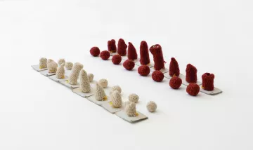 L'image montre une composition artistique composée de petits objets en crochet. Il y a deux groupes disposés sur une base claire : l'un en rouge et l'autre en beige. Les objets de chaque côté varient en forme et en taille, avec des cônes et des boules. La présentation est minimaliste, mettant en avant les textures et les couleurs des créations. L'arrangement est symétrique, créant un effet visuel intéressant.