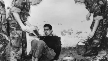 La imagen muestra a un hombre sentado en el suelo, atado y aparentemente en una situación de interrogatorio o detención. Tiene una expresión de tensión en su rostro. Dos personas, vestidas con uniformes militares, están de pie a su alrededor. Una de ellas sostiene un documento y parece que está hablando con el detenido, mientras que la otra sostiene un utensilio, probablemente para tomar notas o hacer preguntas. El ambiente es austero y parece un lugar al aire libre, con una pared desgastada de fondo. La escena capta un momento de tensión y conflicto.