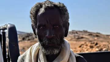الصورة تظهر رجلاً مسنًا ذا لحية وشعر مجعد، يبدو عليه علامات الزمن والخبرة. عينيه تعكسان نظرة واضحة وقوية، بينما يتواجد في خلفية طبيعة جافة تتضمن صخور وتضاريس غير مستوية. يظهر الرجل بملابس بسيطة، مما يعكس بساطة الحياة في المنطقة التي يعيش فيها. السماء زرقاء والجو يبدو صافياً.