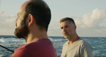 L'image montre deux hommes se tenant près de la mer. L'un d'eux a une barbe et porte un t-shirt rouge, tandis que l'autre a les cheveux grisonnants et un t-shirt clair. L'arrière-plan présente un océan calme sous un ciel bleu parsemé de nuages. L'atmosphère semble sereine, mais les expressions des hommes suggèrent une conversation intense ou réfléchie.