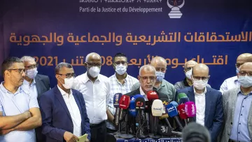 تظهر في الصورة مجموعة من الرجال يقفون أمام لافتة تحمل شعار حزب العدالة والتنمية (Parti de la Justice et du Développement) في المغرب. يتحدث أحدهم، بينما يقف الآخرون خلفه، ومعظمهم يرتدون كمامات. يبدو أن هناك اهتمامًا كبيرًا من وسائل الإعلام، حيث تتواجد عدة ميكروفونات أمامهم. خلفهم، توجد جملة تشير إلى الاستحقاقات التشريعية والاجتماعية. المنظور العام يوحي بحدث رسمي يتعلق بالشؤون السياسية أو الانتخابات.