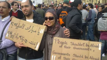L'immagine mostra un gruppo di persone in una manifestazione. Due individui in primo piano tengono in mano dei cartelli. Uno dei cartelli è scritto in arabo e l'altro in inglese. Il messaggio in inglese dice "Le persone non dovrebbero avere paura dei loro governi. I governi dovrebbero avere paura del loro popolo." I partecipanti sembrano impegnati e determinati, con un'ampia folla di persone sullo sfondo. La scena evoca un senso di protesta e richiesta di cambiamento sociale.