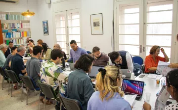 L'image montre une salle de réunion où un groupe de personnes est assis autour d'une grande table. Plusieurs participants semblent travailler sur des ordinateurs portables, tandis que d'autres consultent des documents. Les murs sont décorés de livres et d'illustrations, et plusieurs fenêtres laissent entrer la lumière naturelle. L'ambiance apparaît studieuse et collaborative.