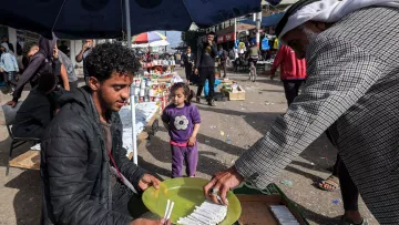 L'immagine mostra una scena di mercato all'aperto, probabilmente in una città. Un giovane uomo è seduto su un contenitore di legno, di fronte a un tavolo, mentre tiene in mano un piatto. Sta vendendo o mostrando dei prodotti, probabilmente cibo o articoli vari. Accanto a lui, un uomo anziano, vestito con indumenti tradizionali, si avvicina per prendere qualcosa dal piatto. Sullo sfondo, ci sono altre persone che passeggiano e alcune bancarelle con vari articoli in esposizione. Una bambina, vestita con una maglietta, osserva curiosa la scena. La luce del giorno illumina vividamente l'ambiente, creando un'atmosfera vivace e animata.