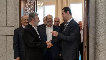 تظهر الصورة مجموعة من الرجال في قاعة استقبال. يتوسط الصورة شخصان يتصافحان، حيث يبدو أن أحدهما في وضعية حديث مع الآخر. الخلفية تحتوي على أبواب وزخرفة رائعة، وهناك أشخاص آخرون يقفون خلفهم، يظهر عليهم تفاعل إيجابي ورغبة في التواصل. الأجواء تبدو رسمية، مما يشير إلى لقاء مهم أو مناسبة خاصة.