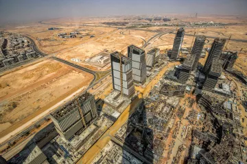 The image showcases a panoramic view of a vast construction site in a desert landscape. In the foreground, several high-rise buildings are partially completed, with cranes and construction equipment evident. The surrounding area features empty land and some roadways, characteristic of a developing urban environment. The backdrop includes a clear sky and distant structures, suggesting a mixture of ongoing urbanization and natural desert terrain. The overall scene illustrates the rapid expansion of urban infrastructure in a typically arid region.