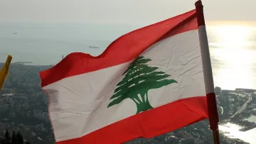 L'image montre le drapeau du Liban flottant au vent, avec un fond qui semble représenter une vue panoramique d'une ville côtière. Le drapeau se compose de trois bandes horizontales : rouge en haut et en bas, et une bande blanche au centre avec un grand cèdre vert, emblème national du Liban. L'horizon présente une mer scintillante, probablement au coucher du soleil, ce qui ajoute une ambiance paisible et naturelle à la scène.