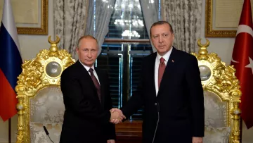 En la imagen se pueden ver a dos líderes políticos, uno de Rusia y otro de Turquía, dándose la mano en un acto formal. Ambos están vestidos con trajes oscuros y corbatas, y parecen estar en un entorno elegante, rodeados de decoraciones doradas y banderas de sus respectivos países. La atmósfera sugiere un momento de colaboración o acuerdo entre las dos naciones.