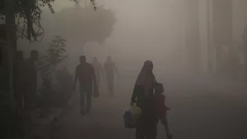L'immagine mostra una scena di una strada avvolta nella nebbia o nella polvere. In primo piano, una donna con un velo scuro tiene la mano di un bambino, mentre camminano insieme. Sullo sfondo, si possono vedere altre persone che camminano, ma sono poco visibili a causa della foschia. Gli alberi e alcune piante ai lati della strada creano un'atmosfera un po' opprimente. La luce sembra attenuata, dando un'impressione di desolazione.