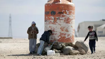 L'image montre un groupe d'enfants autour d'un réservoir d'eau. Le réservoir, de couleur orange et marqué par des signes d'usure, semble abriter l'eau potable. Les enfants, dont l'un est en train de remplir un récipient, sont habillés de manière simple et semblent se trouver dans un environnement désertique. On peut voir des pierres autour du réservoir, et le paysage en arrière-plan est sobre, suggérant une situation de besoin en ressources.