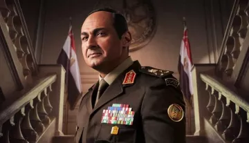 يظهر في الصورة رجل يرتدي زيًا عسكريًا يحمل العديد من الأوسمة والرتب. يبدو أنه يتخذ وضعًا رسميًا وجادًا، مع خلفية تتضمن علمين. تعكس تعابير وجهه الثقة والسلطة، كما أن التصميم العام للصورة يوحي بأجواء عسكرية أو حكومية.