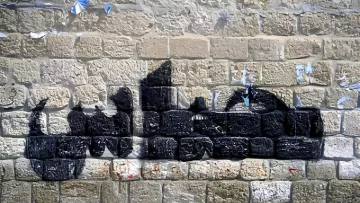 L'immagine mostra un murale su un muro di pietra. Il disegno rappresenta una silhouette di un veicolo, probabilmente un camion, realizzato con vernice nera. Le linee e le forme sono stilizzate, creando un contrasto con la texture della parete di pietra. Sul muro si notano anche resti di manifesti strappati e segni di usura, che aggiungono carattere e storia all'ambiente.