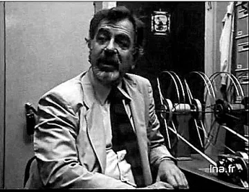 L'image présente un homme assis dans un bureau. Il porte un costume clair et une cravate foncée. Son expression faciale semble animée, presque passionnée, comme s'il était en train de discuter ou d'expliquer quelque chose. À l'arrière-plan, on peut voir des bobines de film ou des documents, ce qui donne une impression d'environnement de travail lié à la production ou à la recherche. L'éclairage est sombre, créant une ambiance vintage, typique des années passées.