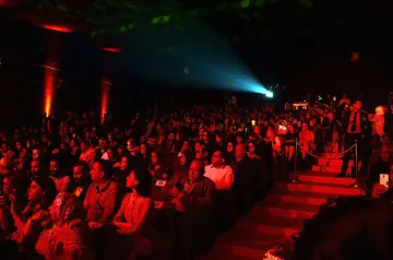 L'image montre un auditorium rempli de spectateurs. La salle est faiblement éclairée avec des lumières rouges qui créent une ambiance chaleureuse et dynamique. Au centre, un projecteur émet un faisceau lumineux, ajoutant à l'atmosphère du moment. Les spectateurs, composés d'hommes et de femmes, semblent attentifs et engagés, certains tenant des téléphones. L'escaliers sur le côté de la scène permet l'accès à d'autres niveaux. L'ensemble de la scène évoque un événement artistique ou culturel.