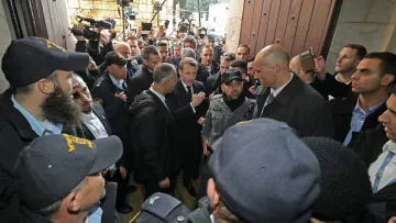 L'image montre une scène animée avec de nombreuses personnes rassemblées autour d'un groupe central. Des agents de sécurité, en uniforme, semblent encadrer des individus, probablement des personnalités importantes. L'atmosphère est tendue, avec des caméras et des journalistes présents, suggérant que cet événement pourrait avoir une importance médiatique. L'ensemble se déroule devant une grande porte, indiquant un lieu significatif, possiblement un bâtiment officiel ou historique.