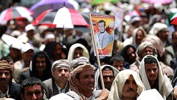 L'image montre une grande foule de personnes réunies, probablement lors d'une manifestation ou d'un rassemblement. La majorité des participants portent des vêtements traditionnels et certains sont abrités sous des parapluies de différentes couleurs. Au premier plan, un homme tient une affiche avec un portrait, ce qui suggère qu'il soutient une personnalité ou une cause spécifique. L'atmosphère semble engagée et collective.